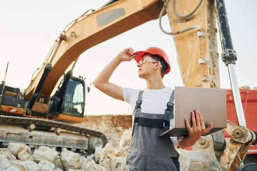 Standing against yellow vehicle. Worker in professional uniform is on the borrow pit at daytime.