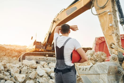 Standing against yellow vehicle. Worker in professional uniform is on the borrow pit at daytime.