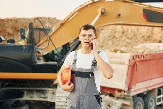 Standing against yellow vehicle. Worker in professional uniform is on the borrow pit at daytime.