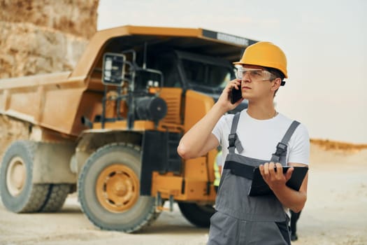 In glasses and hard hat. Worker in professional uniform is on the borrow pit at daytime.
