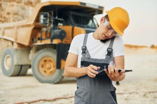 In glasses and hard hat. Worker in professional uniform is on the borrow pit at daytime.