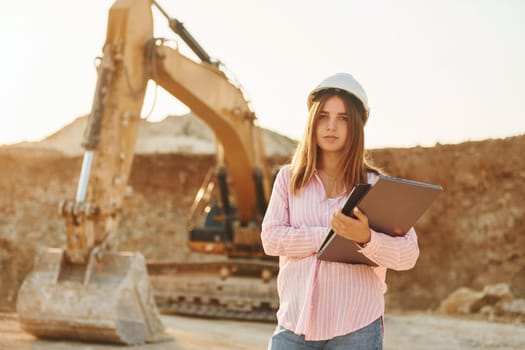 With documents. Young woman with documents in hard hat is standing on the borrow pit.