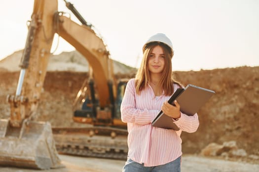 With documents. Young woman with documents in hard hat is standing on the borrow pit.