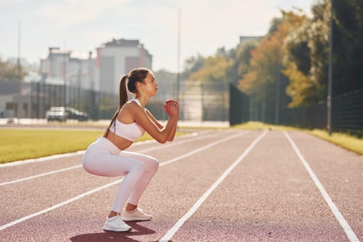 Doing squats. Young woman in sportive clothes is exercising outdoors.