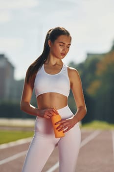 Holding bottle of watter. Young woman in sportive clothes is exercising outdoors.