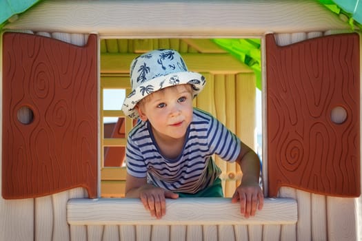 A cute little boy in a sailor's shirt looks out the window of a toy house in surprise