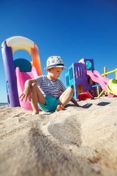 A little boy in a hat sits on a sand and play with it, beach and colourful playground in a background. A beautiful summer day, free time and a carefree childhood