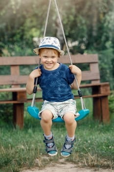 Happy little boy with hat having fun on swing in domestic garden. Healthy toddler child swinging on sunny summer day. Children activity outdoor, active smiling kid laughing.