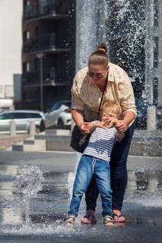 Mom with her son in the city by the water fountain. The little child is frightened by the sudden jets of water. Kids fun in the city on a hot summer day.