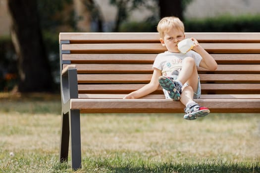 Little boy with is sitting on bench in park and drinks juice from plastic cup.