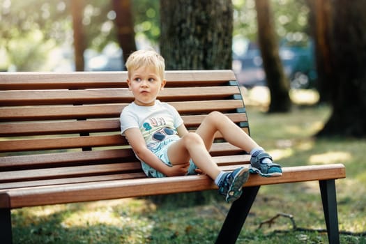 Cute kid boy sitting on a bench in the park and rest.