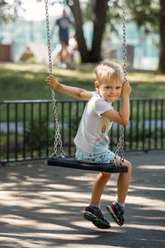 A mischievous little boy swings in a park on a chain swing, the child turns to the camera and smiles happily.