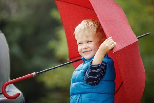 Adorable little boy with red umbrella and blue jacket outdoors at rainy day.