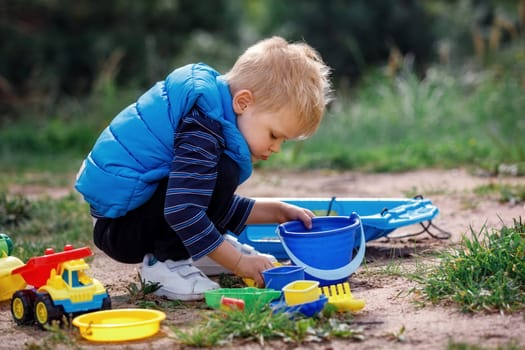 Child squat on the ground playing with sand toys. The child digs the sand into a plastic bucket. Summer outdoor activity for kids. Leisure Time.