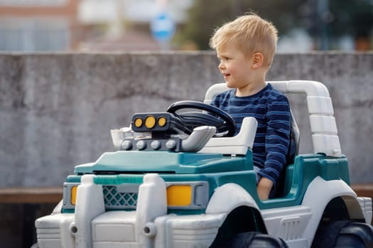 Happy blond little boy in a big toy battery powered car outdoors.