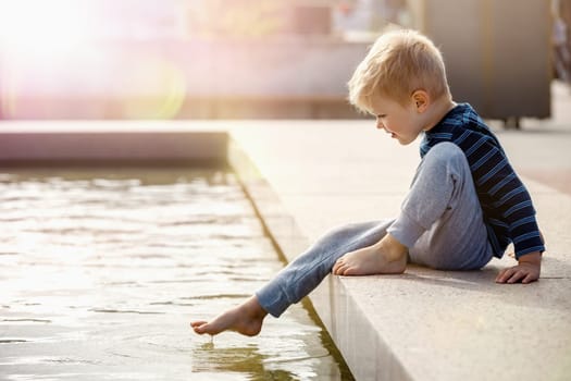 A curious child, a boy, barefoot touches the water of a city fountain. Hot and sunny summer day, the water is a good refreshment for the boy's feet.
