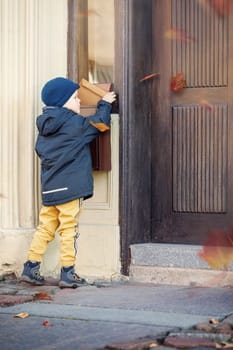 Cute boy sending or receiving mail with mailbox near house. Autumn, the wind blows colorful tree leaves.