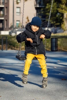 A cute boy in warm autumn clothes, yellow pants and a blue hat swings on a chain swing with a smile.