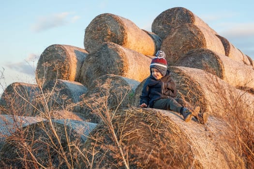 A child wearing warm autumn clothes and a hat sits in the evening light and plays on a pile of golden straw rolls in the village.