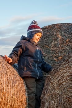 A cute boy poses on top of a pile of straw bales, sunset lights, a background of blue sky.