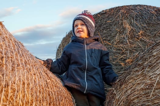 A smiling cute boy in a knitted hat stands on a golden stack of straw in the evening sun. The child's face is illuminated by the evening sun, he is climbed high, and looks into the distance.