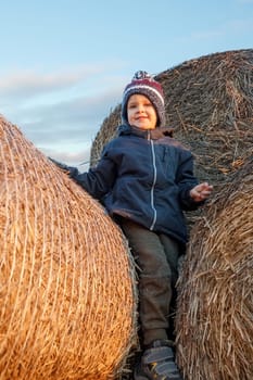 A smiling cute boy with a knitted hat stands on a golden straw stack in the evening sunlight.