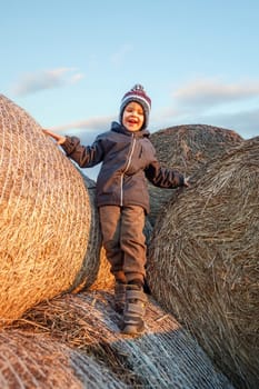 A smiling cute boy with a knitted hat stands on hay bales in the evening sunlight.