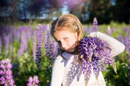 portrait of cute little happy two year old kid girl with bloom flowers lupines in field of purple flowers. Child in nature concept.