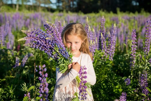portrait of cute little happy two year old kid girl with bloom flowers lupines in field of purple flowers. Child in nature concept.