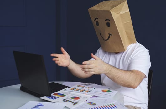 A man is a businessman in a white shirt with a paper bag on his head, with a joyful smiley face drawn, conducts a video conference or training via video link on a laptop. Emotions and gestures.