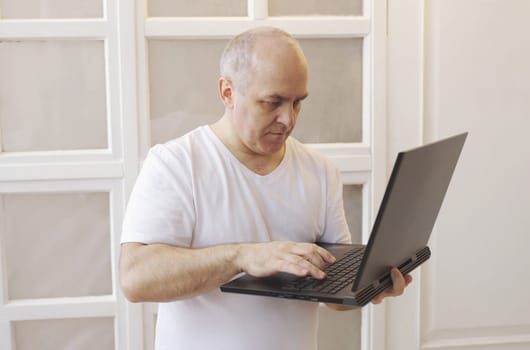 A man holds a laptop in his hands, stands at home and communicates in social networks, writes a message.