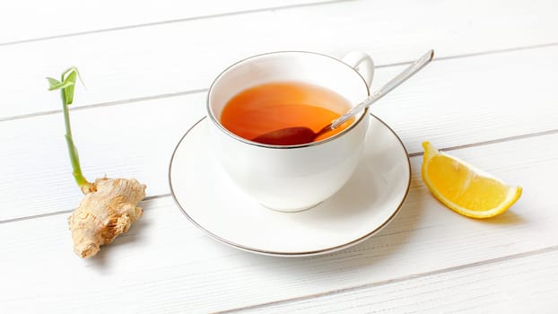 White porcelain cup with hot amber tea, silver spoon in it, dry ginger root green sprout and piece of lemon at side.