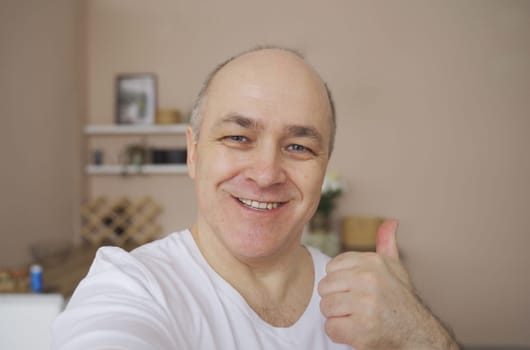 Smiling male blogger taking selfie or video call from his apartment, showing hand gesture.