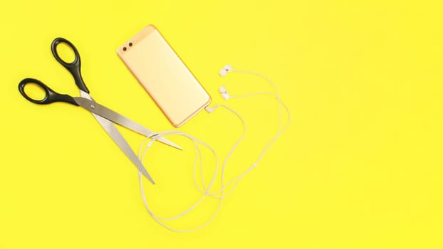 Top view on rose gold cell phone with ear buds cable to be cut with large scissors on yellow background.