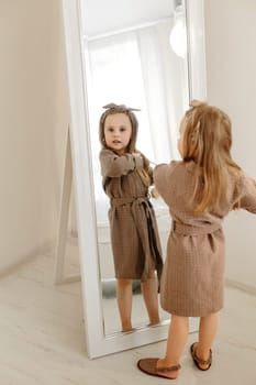 A girl in a brown coat looks through a large mirror at the camera.