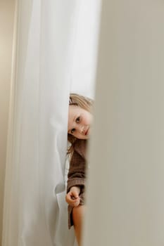 A girl in a brown bathrobe sits on the windowsill and peeps through the curtain, smiling.