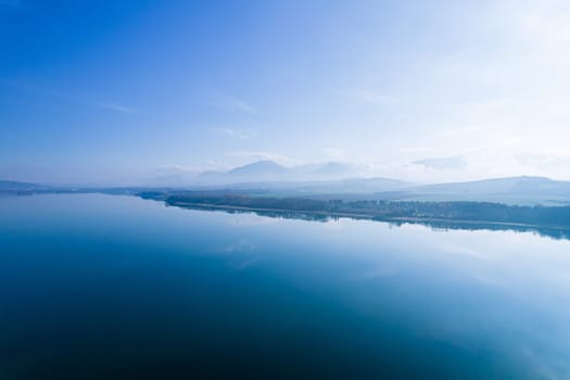 Blue lake and morning fog over forests and mountains. A panoramic view from the top of beautiful nature.