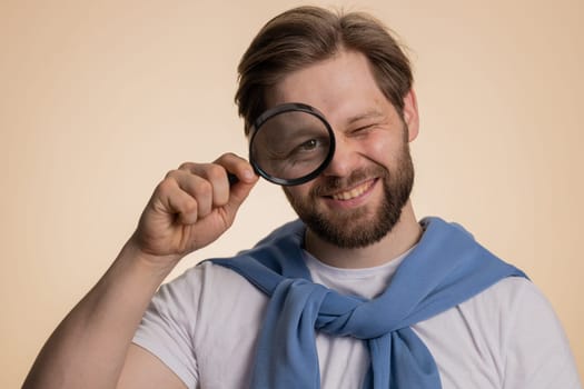 Investigator researcher scientist man holding magnifying glass near face looking into camera with big zoomed funny eyes, searching analysing. Excited caucasian guy isolated on beige studio background