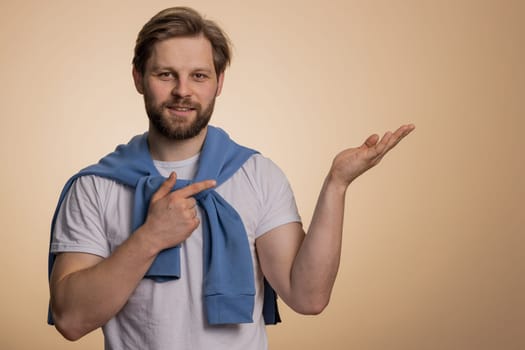 Caucasian man showing thumbs up and pointing empty place, advertising area for commercial text, banner, logo, copy space for goods promotion. Young bearded guy isolated on beige studio background