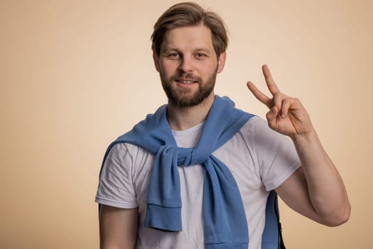 Young caucasian man showing victory sign, hoping for success and win, doing peace gesture, smiling with kind optimistic expression. Handsome bearded guy isolated alone on beige studio background