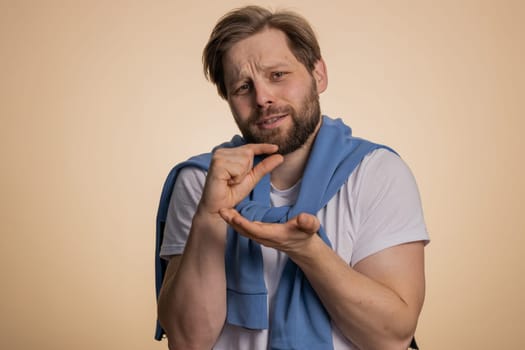 Need some more, please give me. Caucasian man showing a little bit gesture with sceptic smile, showing minimum sign, measuring small size. Handsome guy isolated alone on beige studio background