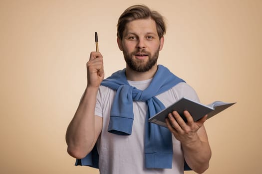 Thoughtful journalist caucasian man making notes, writing down thoughts with pen into notepad notebook diary, to do list, good idea. Handsome bearded guy isolated alone on beige studio background
