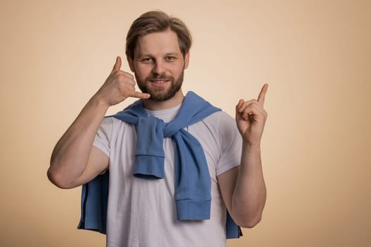 Call us, here is contact number. Handsome bearded caucasian man in t-shirt looking at camera doing phone gesture like says hey you call me back conversation. Guy isolated on beige background, indoor
