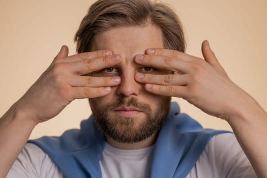 Nosy curious caucasian man closing eyes with hand and spying through fingers, hiding and peeping, binocular gesture, exploring way, seeking something in distance. Young guy on beige studio background