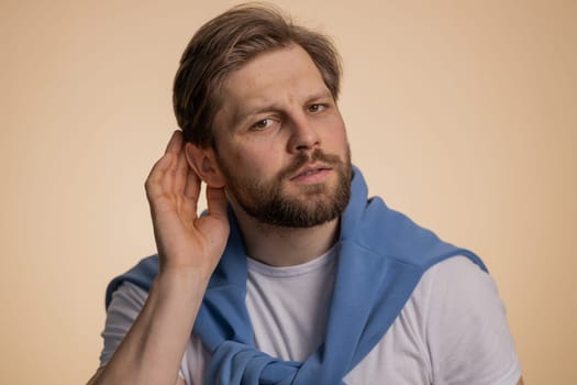 I cant hear you. What. Man trying hear you, looking confused and frowning, keeping arm near ear for louder voice, asking to repeat to hear information, deafness. Young guy isolated on beige background