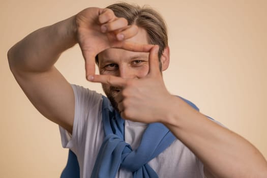 Man photographer gesturing picture frame with hands, looks through fingers and focusing on interesting moment, imitating zoom and cropping nice image. Young guy isolated on beige studio background