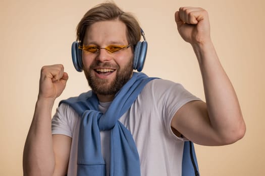 Young happy man listening music via headphones and dancing disco fooling around having fun expressive gesticulating hands, celebrating. Handsome winner guy isolated on beige studio background indoors