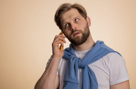 Disinterested young Caucasian man having annoyed boring talk on smartphone. Tired sleepy adult guy talking on mobile phone with friend, making online conversation isolated on studio beige background