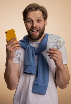 Happy man looking smartphone display sincerely rejoicing win, celebrating, receiving money dollar cash banknotes, success lottery jackpot luck. Young bearded guy isolated on beige studio background