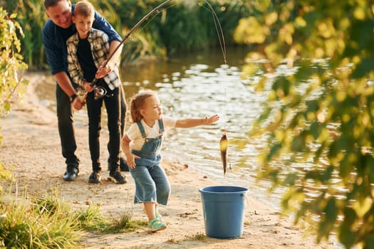 Girl putting fish into the bucket. Father with son and daughter on fishing together outdoors at summertime.
